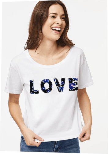 A model wears the Brit Love Tee in White Multi. Hand-cut block letters on the front of the t-shirt spell out the word, Love, across the chest in upcycled Alivia Poppy Play Black. The boxy, cropped tee falls just below the waistband for a loose fit. The wide scooped neckline and drop shoulder short sleeves create an easy pullover.