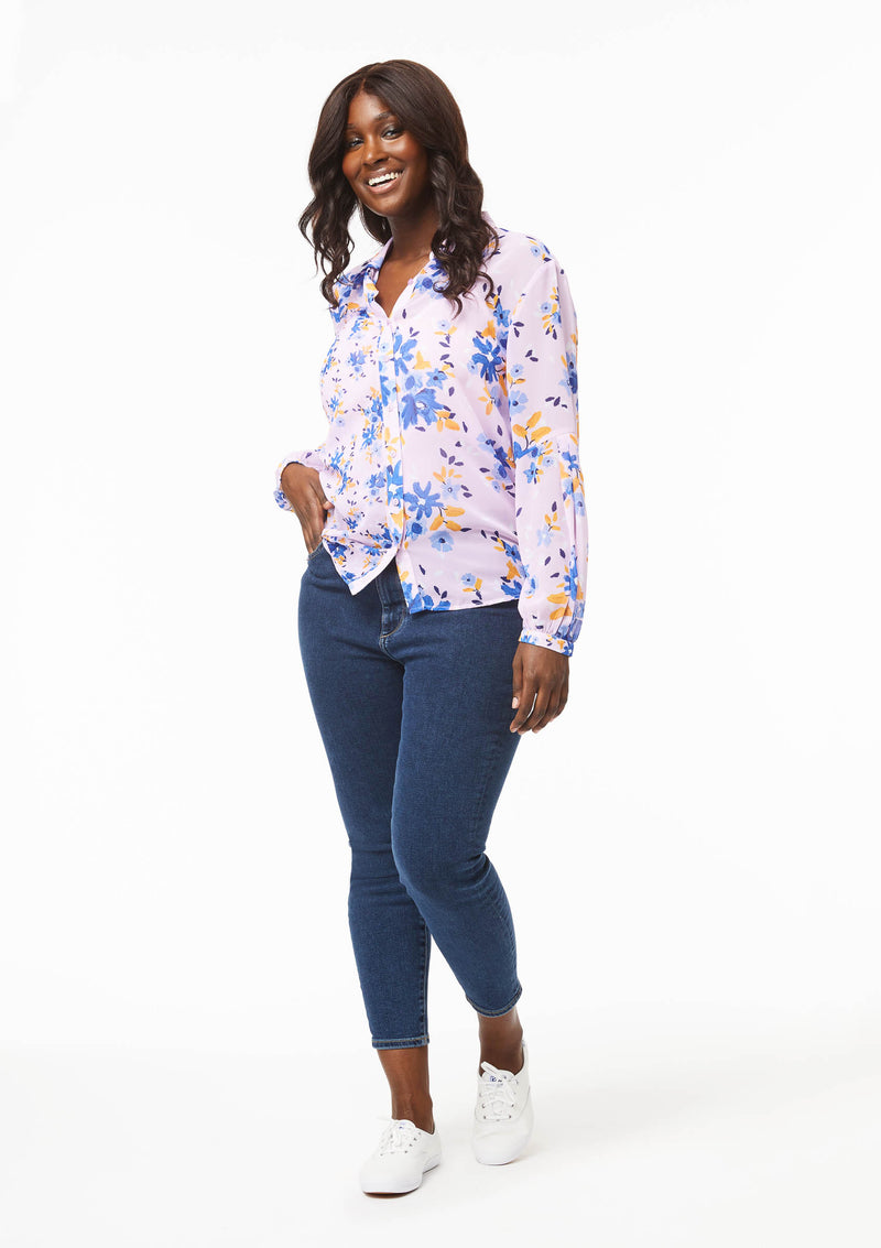 A model with medium dark skin models the Vicki Shirt in Wildflower Burst Orchid paired with solid blue mid-calf jeans and white lace-up canvas shoes. The model has one hand in the pocket of their jeans, pulling up the right panel of the shirt at the hip.