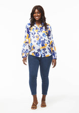 Load image into Gallery viewer, A person with a curvy figure models the Whimsical Floral Vicki Shirt with ankle-length medium blue denim jeans and neutral strappy flats. The hem of the shirt scoops down slightly from the hip, creating a subtle gap between the shirt panels below the bottom button.