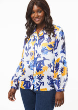 Load image into Gallery viewer, A person with a medium dark skin tone models the Vicki Shirt while standing in counterpoise. The yellows, blues and violets in the fabric are bold and vibrant. Darker blue cuffs are accent the main fabric. The shirt is loose fitting but slimming and paired with a white camisole undershirt and medium blue denim jeans.