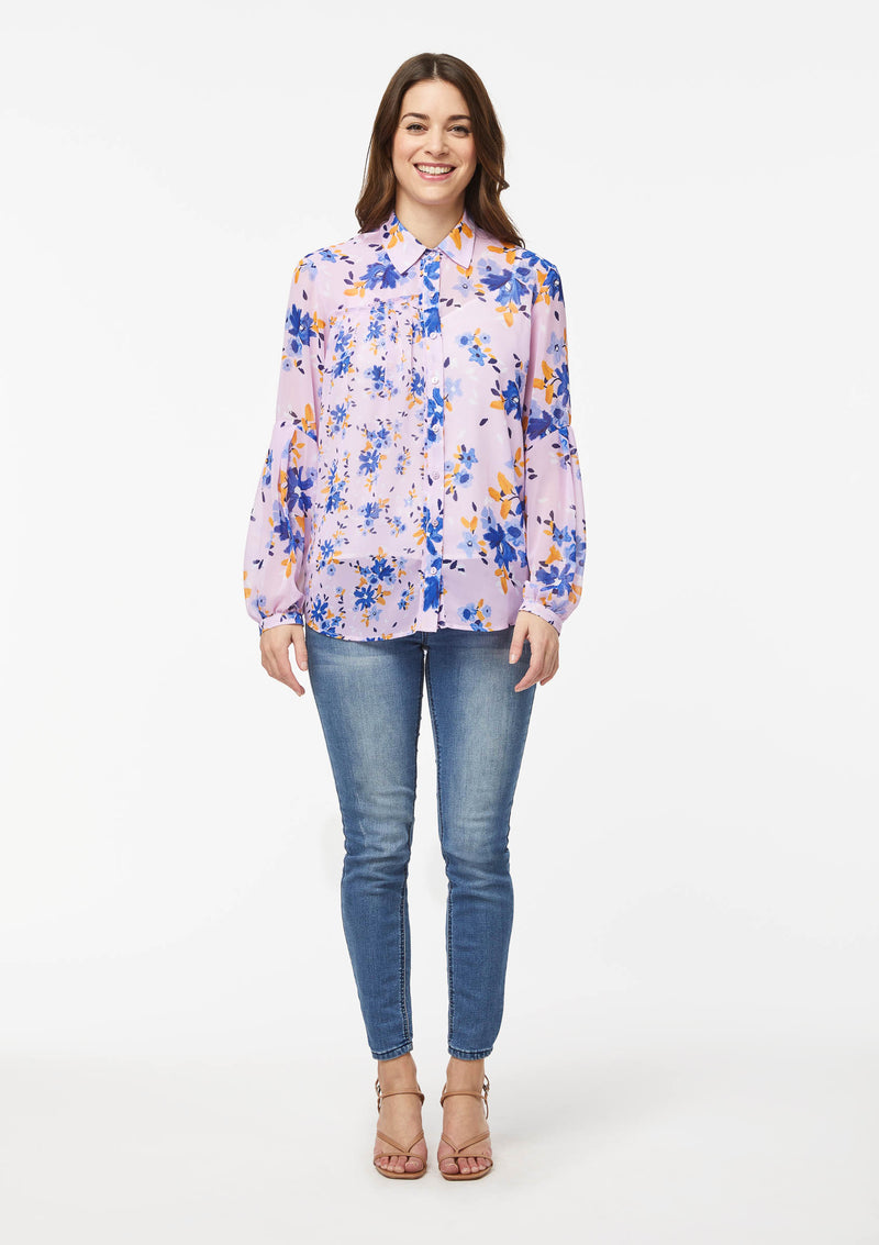 Front view. Alivia’s Vicki Shirt in Wildflower Burst Orchid. A model with light skin shows the shirt paired with light blue skinny jeans and brown strappy heels. The fabric is pale pink with scattered light and dark blue and canary yellow wildflowers. The blue flowers compliment the blue jeans.