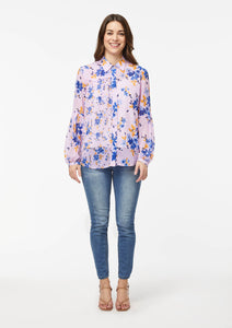 Front view. Alivia’s Vicki Shirt in Wildflower Burst Orchid. A model with light skin shows the shirt paired with light blue skinny jeans and brown strappy heels. The fabric is pale pink with scattered light and dark blue and canary yellow wildflowers. The blue flowers compliment the blue jeans.