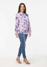 Load image into Gallery viewer, Front side view of the Wildflower Burst Orchid Vicki Shirt. The model’s shoulder and arm are pulled back, revealing a scalloped hem detail at the hip. The hem of the shirt scoops down towards the shirt front and back. With arms resting at the sides, the lower blousonsleeve falls straight down from pleats that begin at the elbow.