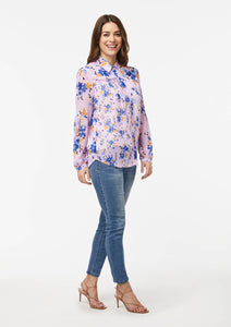 Front side view of the Wildflower Burst Orchid Vicki Shirt. The model’s shoulder and arm are pulled back, revealing a scalloped hem detail at the hip. The hem of the shirt scoops down towards the shirt front and back. With arms resting at the sides, the lower blousonsleeve falls straight down from pleats that begin at the elbow.