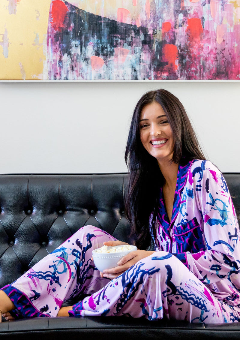 A person wearing the Sofia Pajama Set sits with feet up on a black leather couch, smiling at the camera and holding a bowl of popcorn. The colors in the modern painting behind them complement the Alivia Happy Strokes Pink fabric, draping naturally from their body for comfort and style.