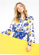 Load image into Gallery viewer, A person with a light skin tone and blonde hair holds a yellow sign while modeling the front of the Vicki Shirt in Alivia’s Whimsical Floral print. The background of the fabric is white with yellow flowers accentuating an abstract floral design of light and dark blue stems and violet flowers with pale pink centers.