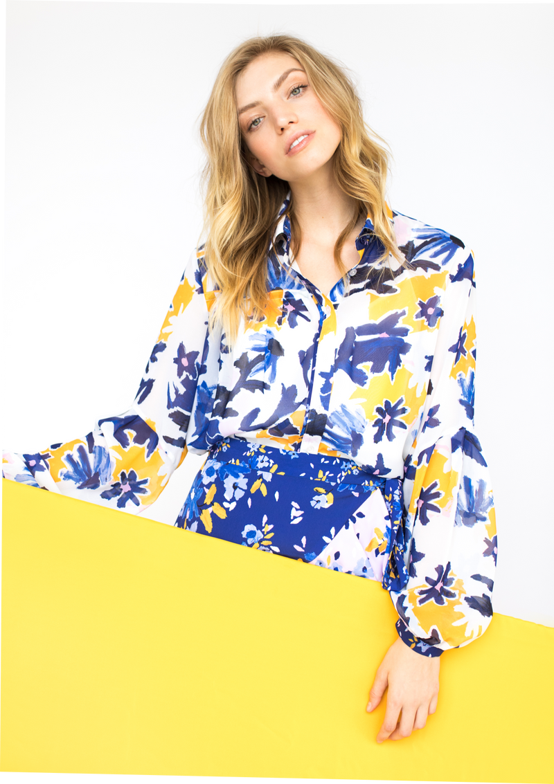 A person with a light skin tone and blonde hair holds a yellow sign while modeling the front of the Vicki Shirt in Alivia’s Whimsical Floral print. The background of the fabric is white with yellow flowers accentuating an abstract floral design of light and dark blue stems and violet flowers with pale pink centers.