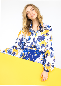 A person with a light skin tone and blonde hair holds a yellow sign while modeling the front of the Vicki Shirt in Alivia’s Whimsical Floral print. The background of the fabric is white with yellow flowers accentuating an abstract floral design of light and dark blue stems and violet flowers with pale pink centers.