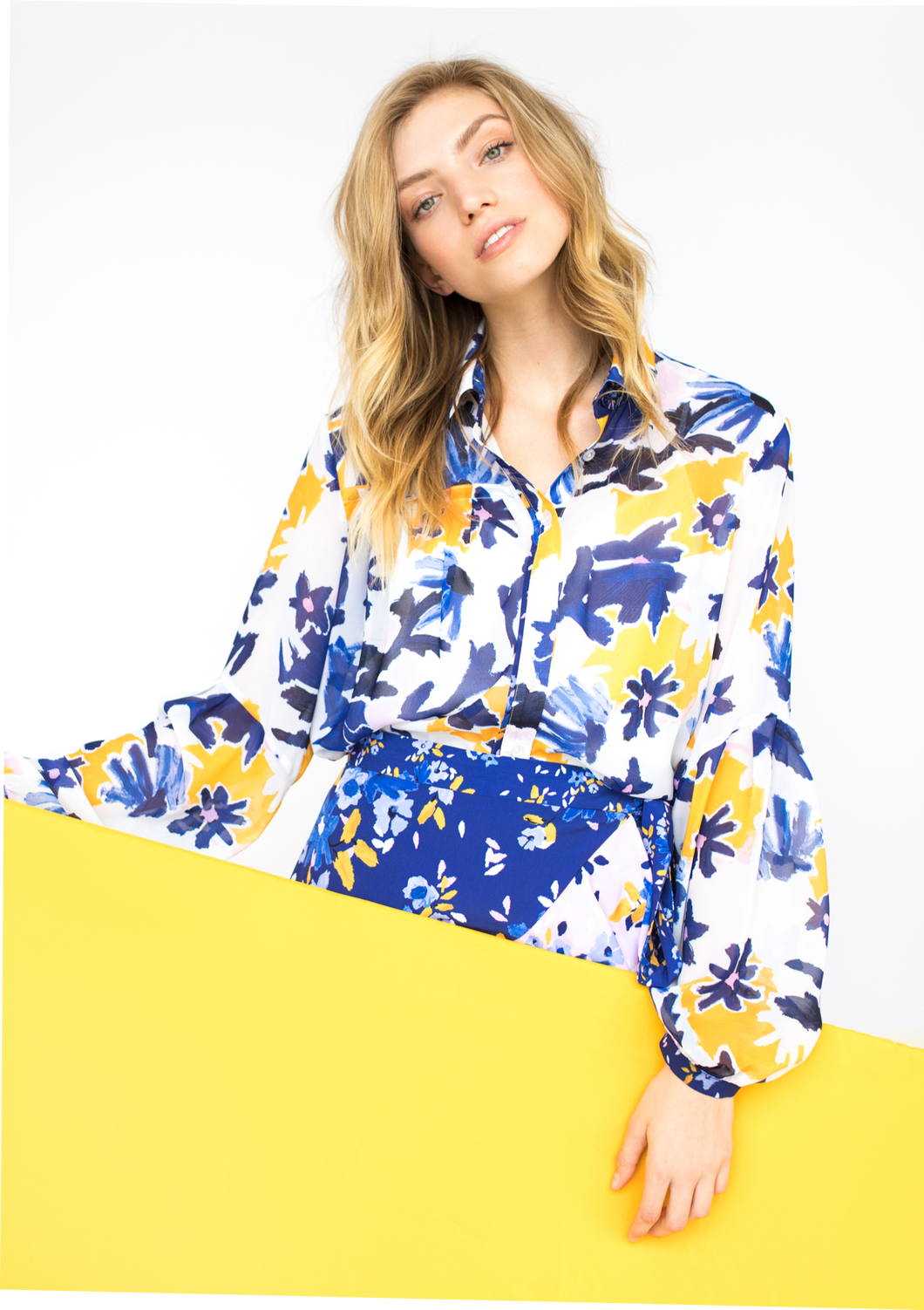 A person with a light skin tone and blonde hair holds a yellow sign while modeling the front of the Vicki Shirt in Alivia’s Whimsical Floral print. The background of the fabric is white with yellow flowers accentuating an abstract floral design of light and dark blue stems and violet flowers with pale pink centers.